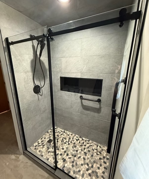 Walk-in shower with gray tile walls, a pebble tile floor and a black-framed glass door