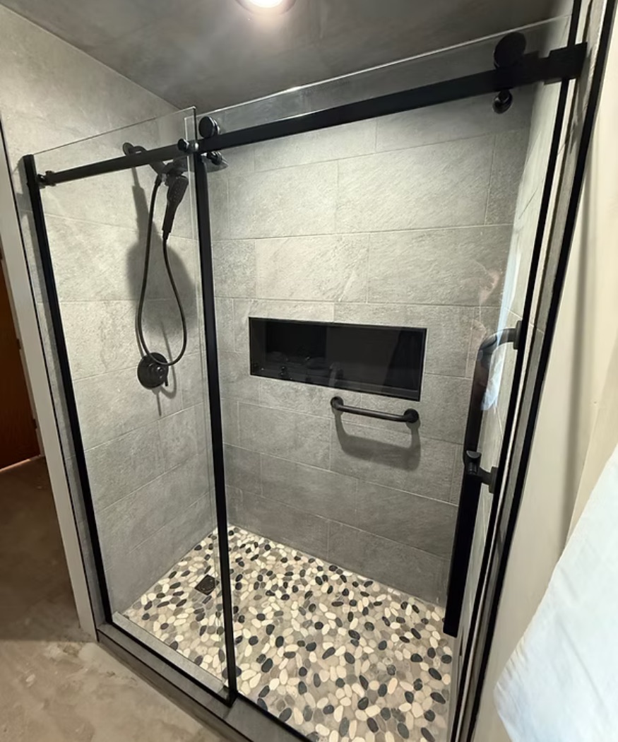 Walk-in shower with gray tile walls, a pebble tile floor and a black-framed glass door